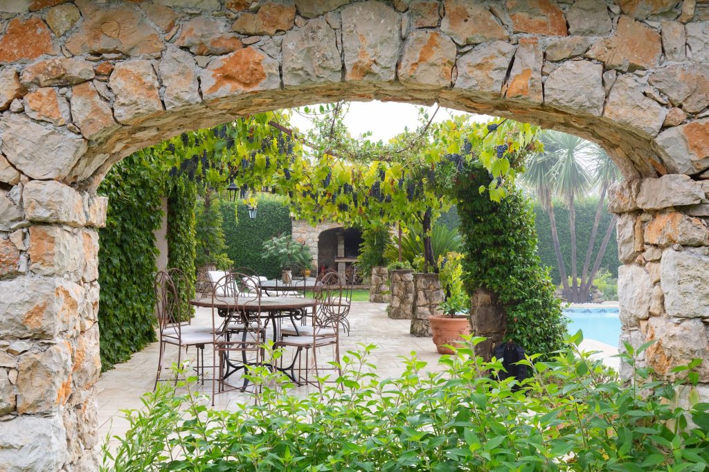 Terrasse paysagée avec pergola végétalisée, murs en pierre et palmiers dans un jardin méditerranéen à Grasse